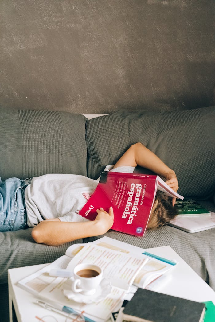 A tired student sleeps on a couch with a book covering their face, surrounded by study materials and coffee.